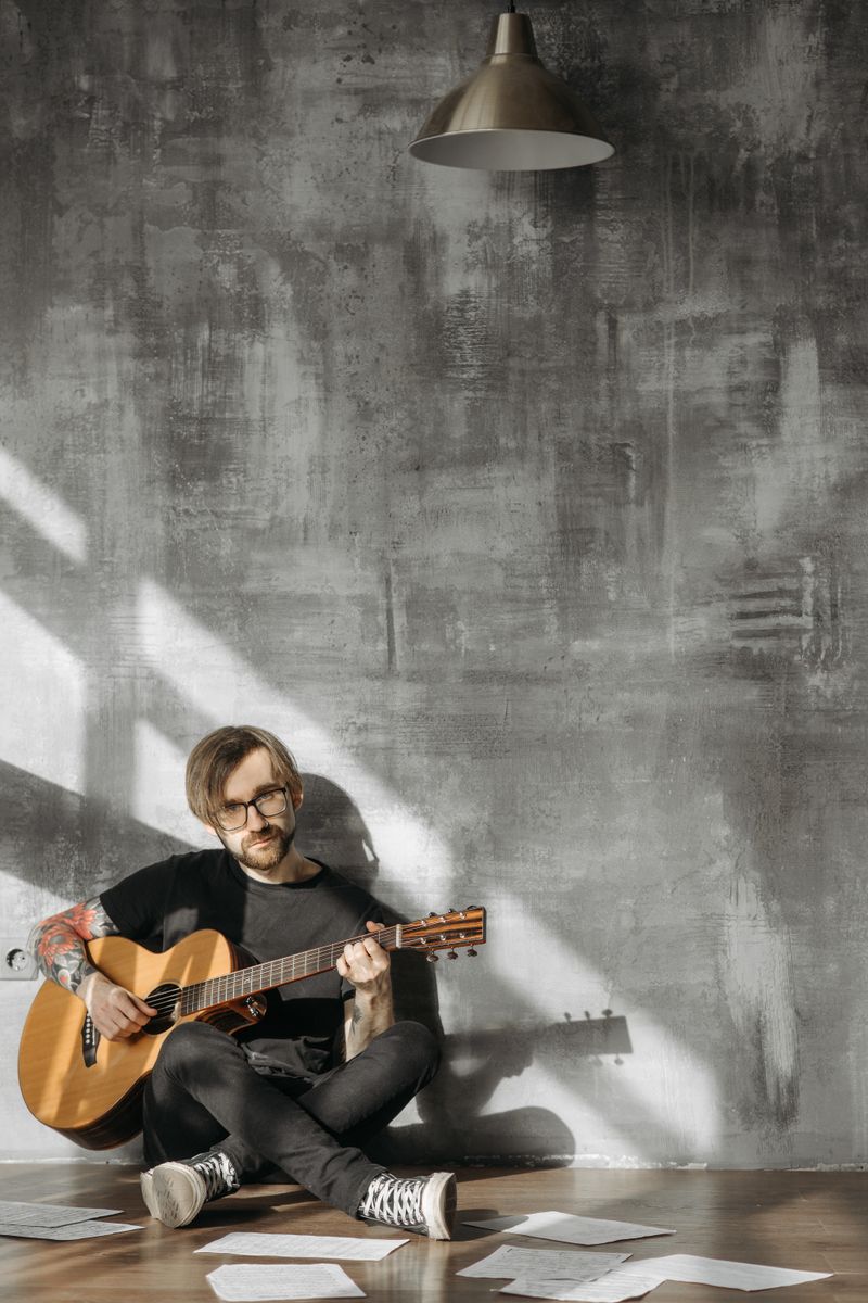 A guitar teacher sitting on the floor playing an acoustic guitar with sheet music scattered around